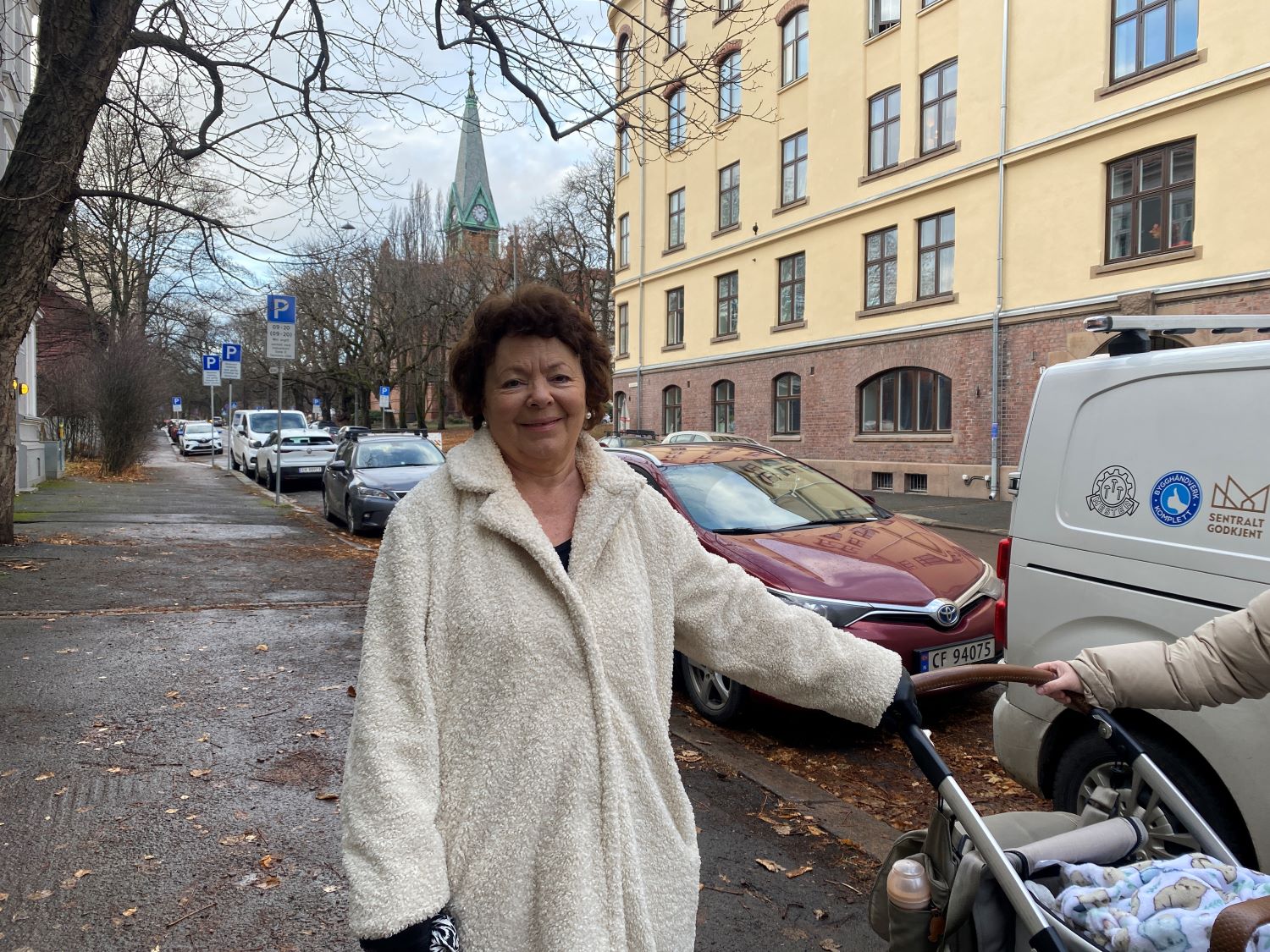 Dame med hvit kåpe på fortauet. Sagene kirke er så vidt i bakgrunnen og hun holder på en barnevogn med høyre arm.