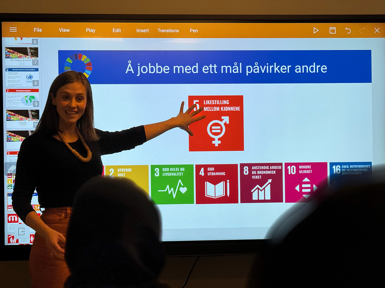 Dame står og holder presentasjon om FNs-bærekraftsmål på en smartboard. Hun har på seg mørk genser og brune bukser.