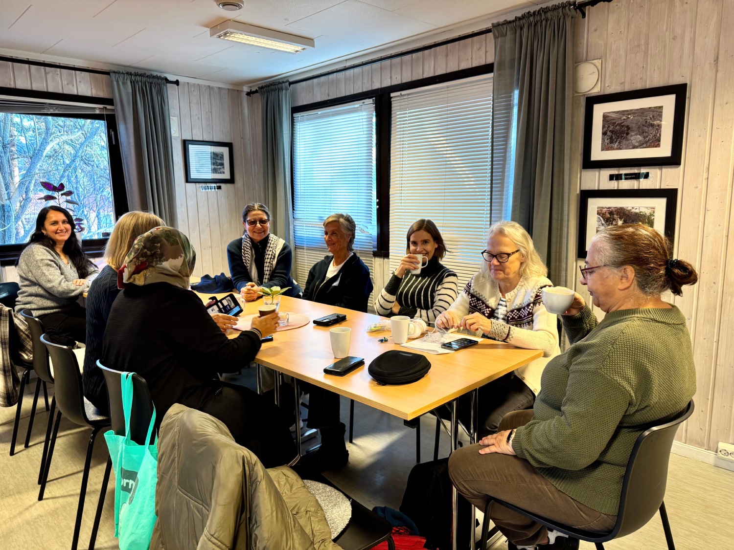 Åtte damer sitter rundt et langbord. En av dem sitter og strikker. Det er flere kaffekopper på bordet. De smiler til kameraet. 