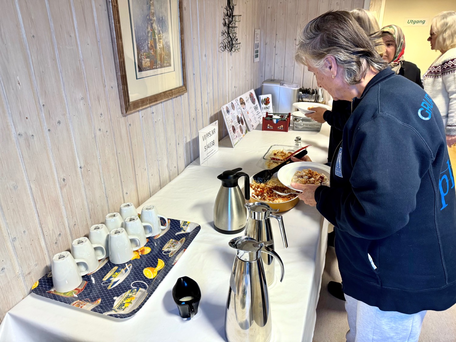 Flere damer som står i kø for å forsyne seg med maten som står på bordet foran seg. På bordet står det også kaffekopper og mugger. 