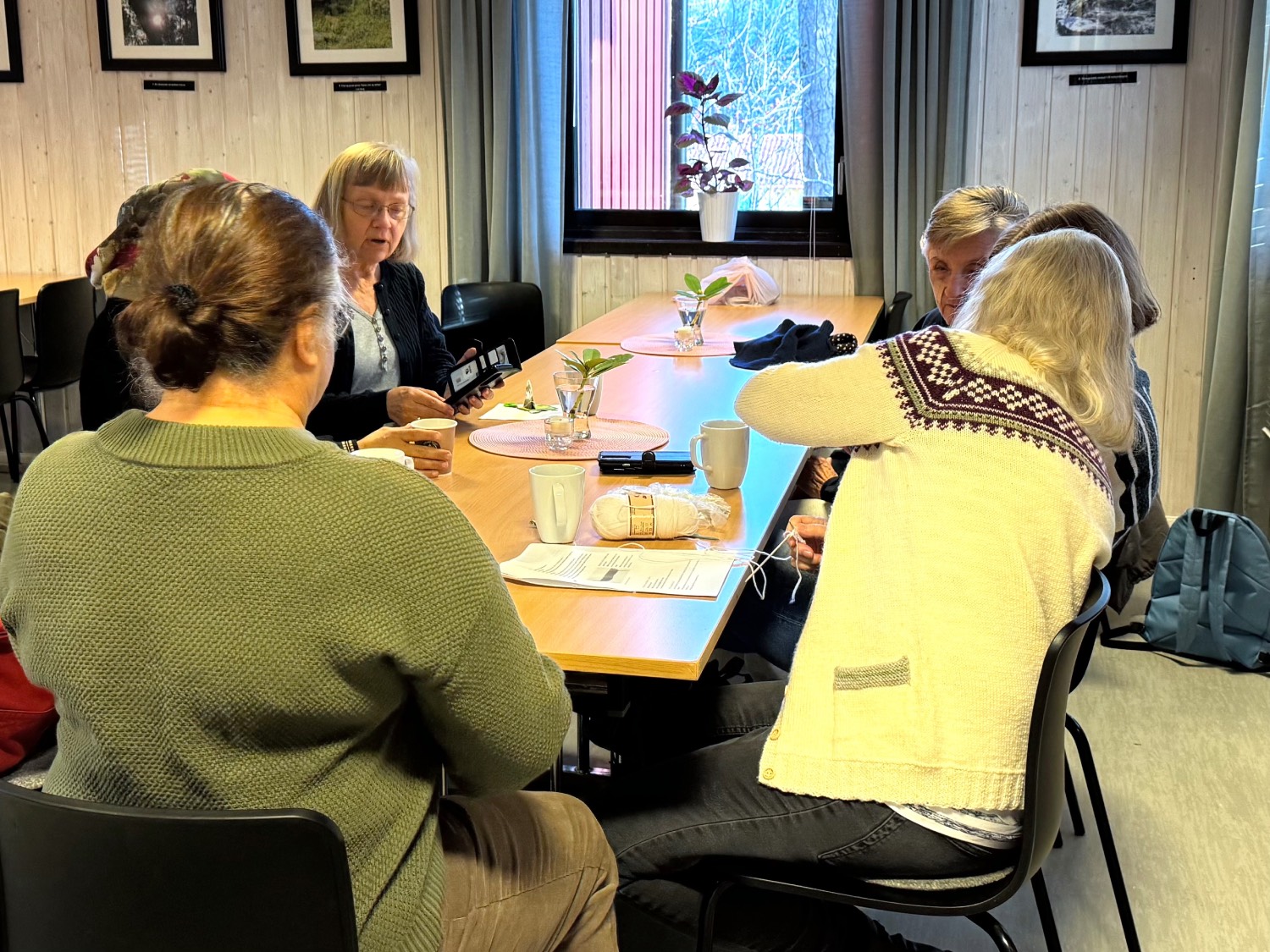 Seks damer som sitter rundt et langbord og prater med hverandre. En har strikketøyet klart på bordet, resten drikker kaffe. 