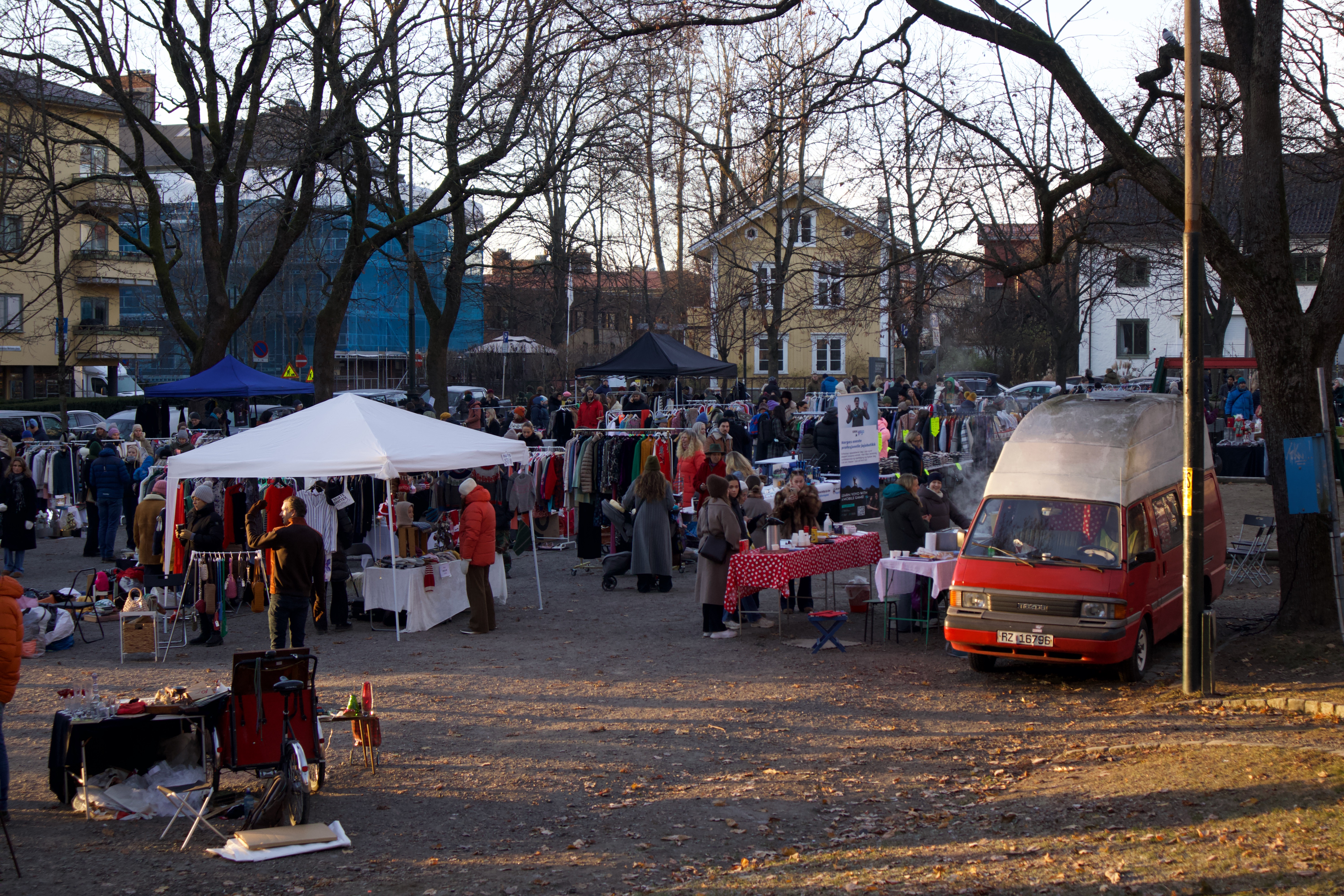 Markedsboder er plassert på en stor grusplass. Mellom dem går det mange mennesker. Til høyre i bildet er det en rød van