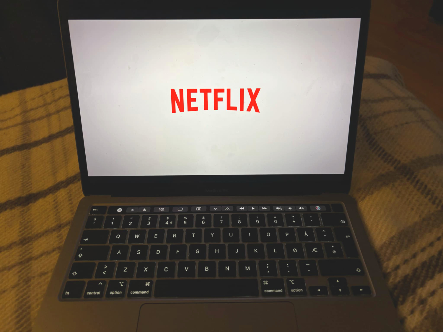 En MacBook med Netflixlogoen på skjermen. Macen er plassert på et rutete teppe, i et mørkt rom.