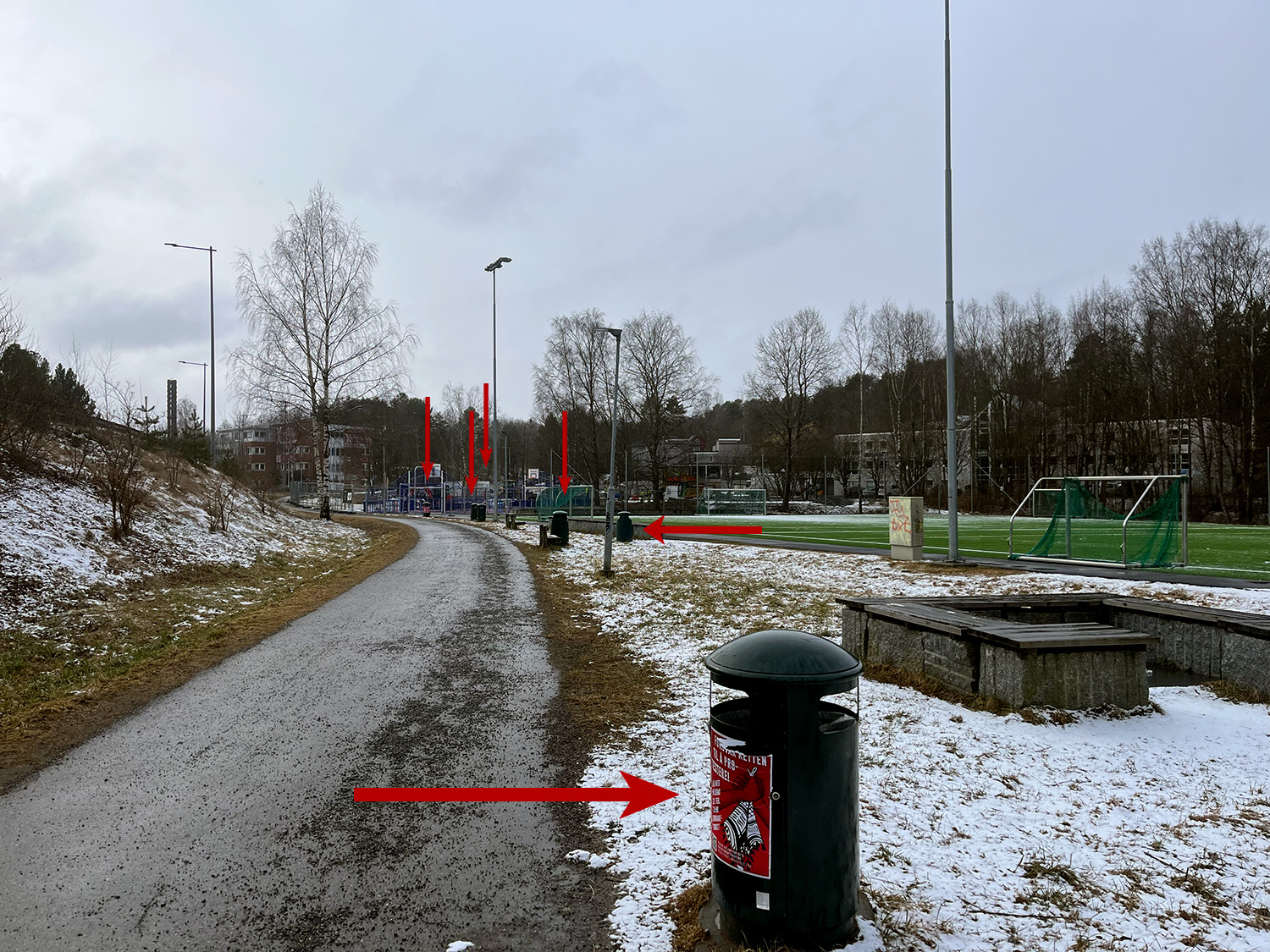 Det står 6 søppelbøtter på rekke ved siden av en liten fotballbane