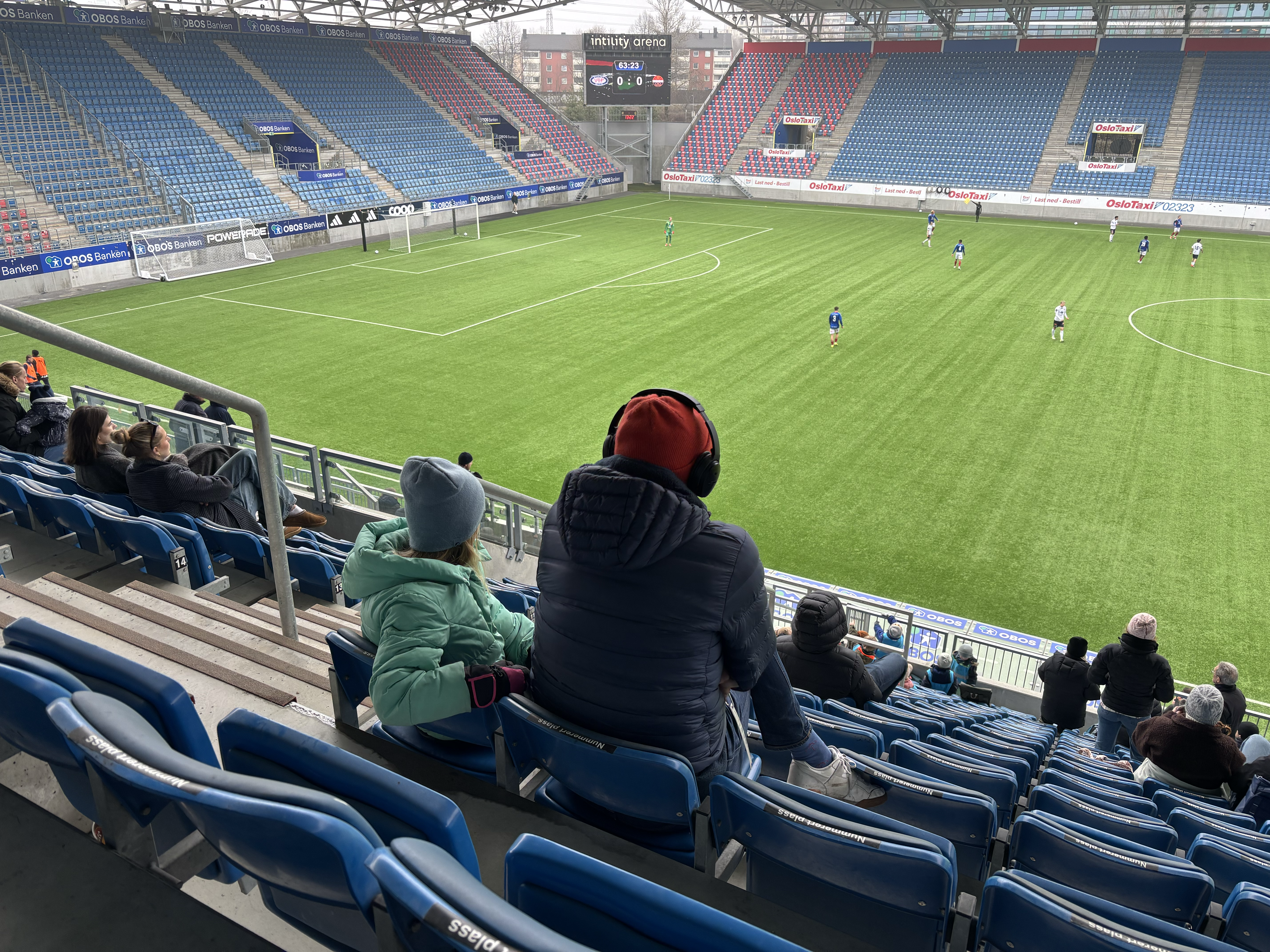 Far og datter ser på fotballkamp, far ser på telefonen, datter følger med på kampen