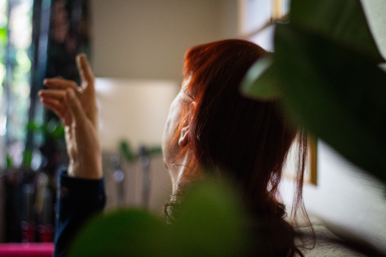 Bilde av en eldre dame som sitter i hjemmet sitt. Hun har løftet en arm og har blikket vendt vekk fra kamera. I forgrunnen ser man noen grønne planter.