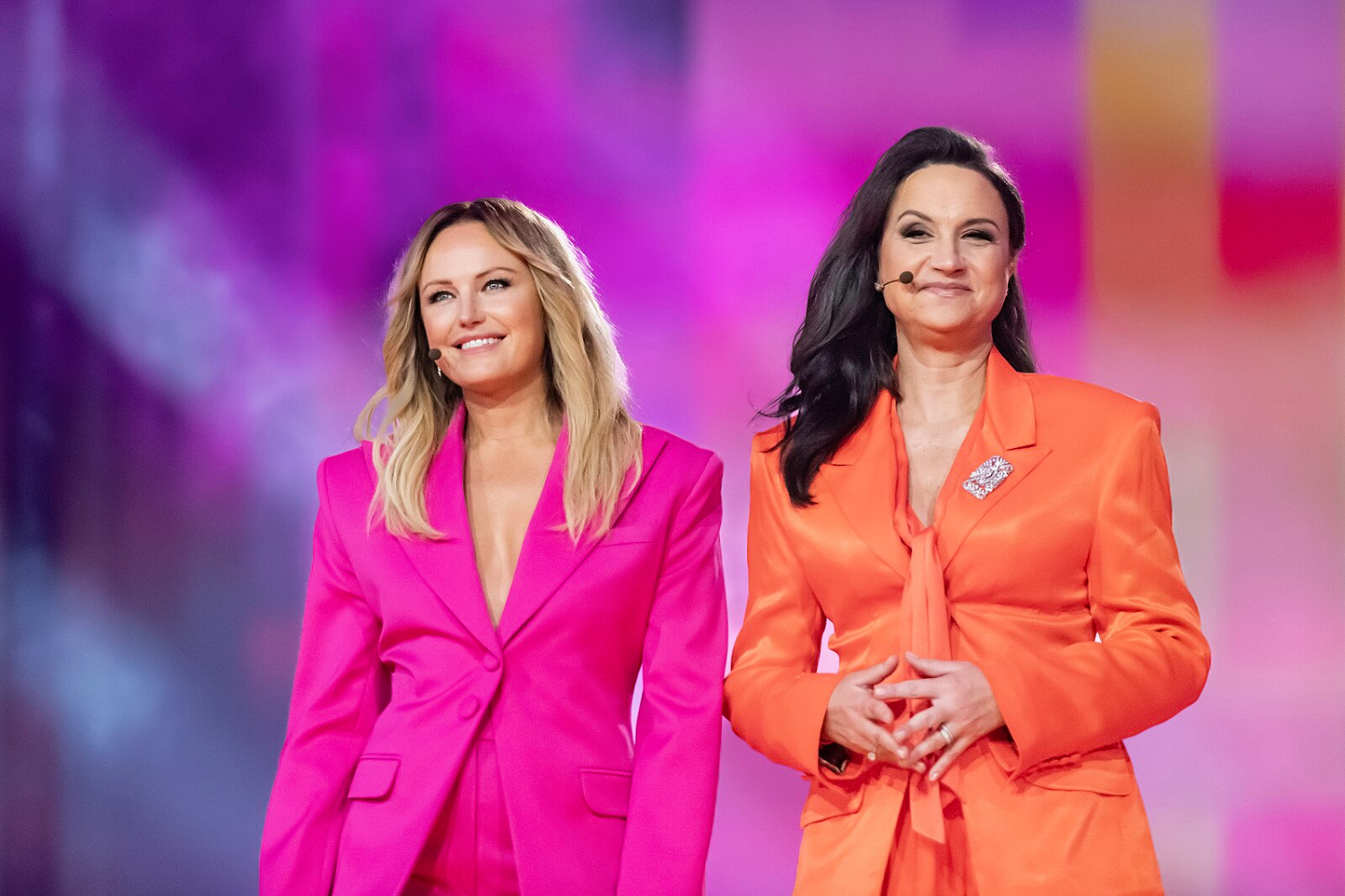 To damer, hun ene er ikledd en rosa dress og hun andre er ikledd en oransje dress. De to damene stor foran en fargerik bakgrunn.