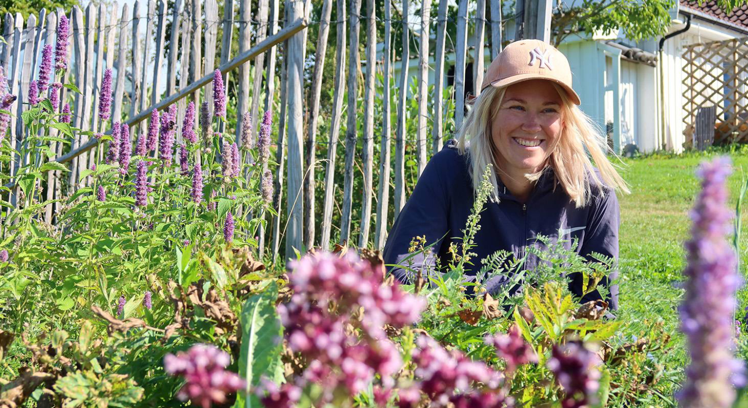 Bonde Marit sitter på huk bak en lilla blomstereng, og smiler mot solen.