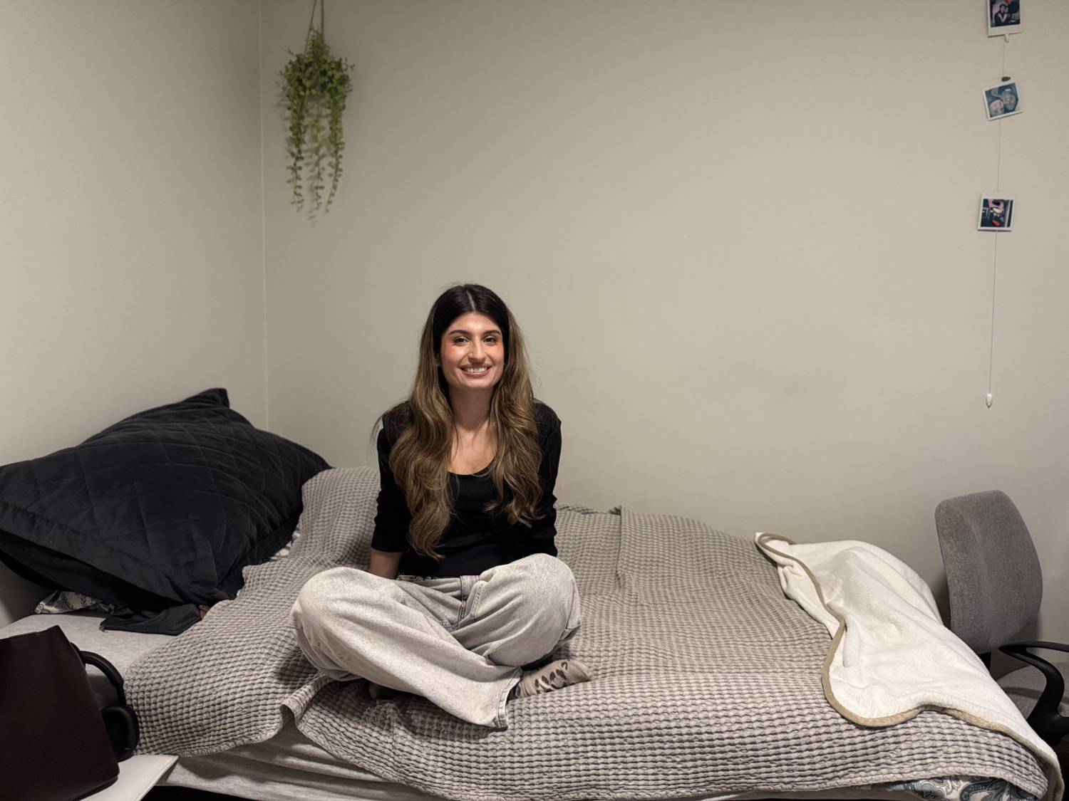 ung dame med langt bølgete mørkt hår sitter med begna i kryss på en seng. veggen bak og dyna på senga er i en lys grå farge