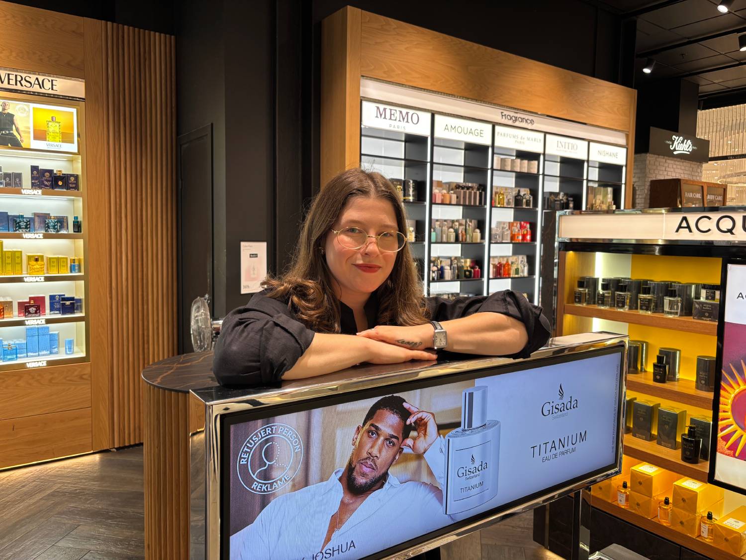 ung dame med lyst brunt hår og briller lener seg over en reklameplakat for en ny herreparfyme. i bakgrunnen er det hyller med mange forskjellige parfymeflasker