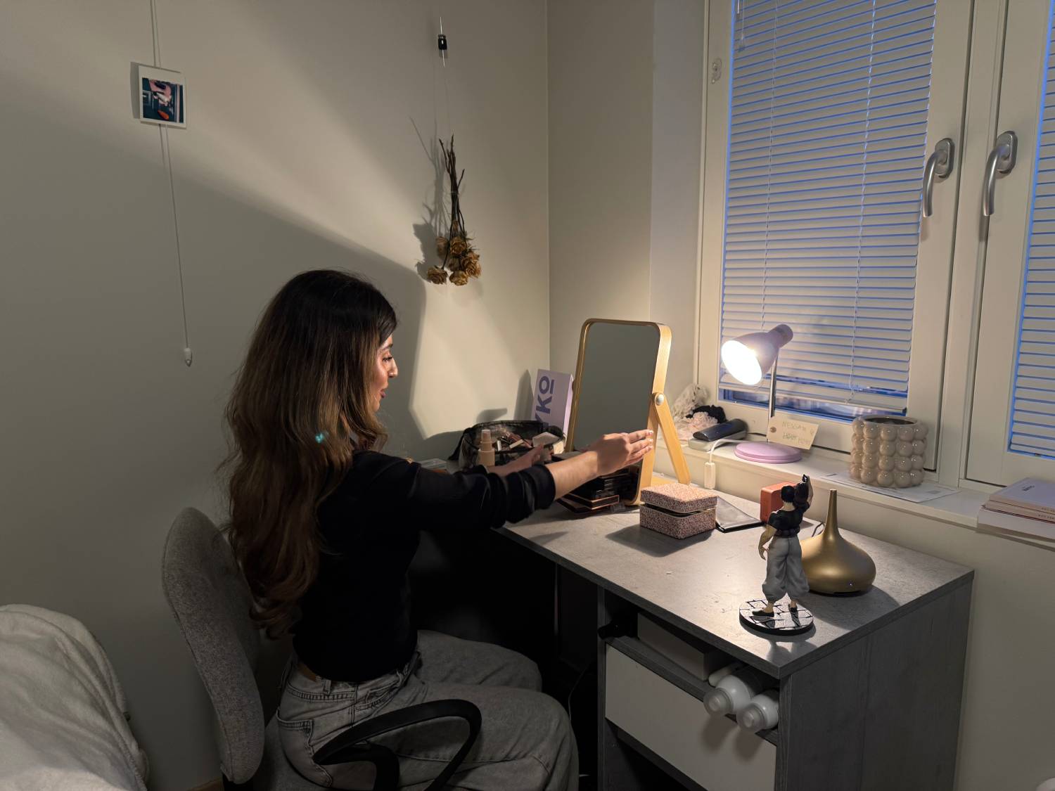 jente i svart genser sitter med ryggen til kamera foran en grå pult med et lite sminke speil på