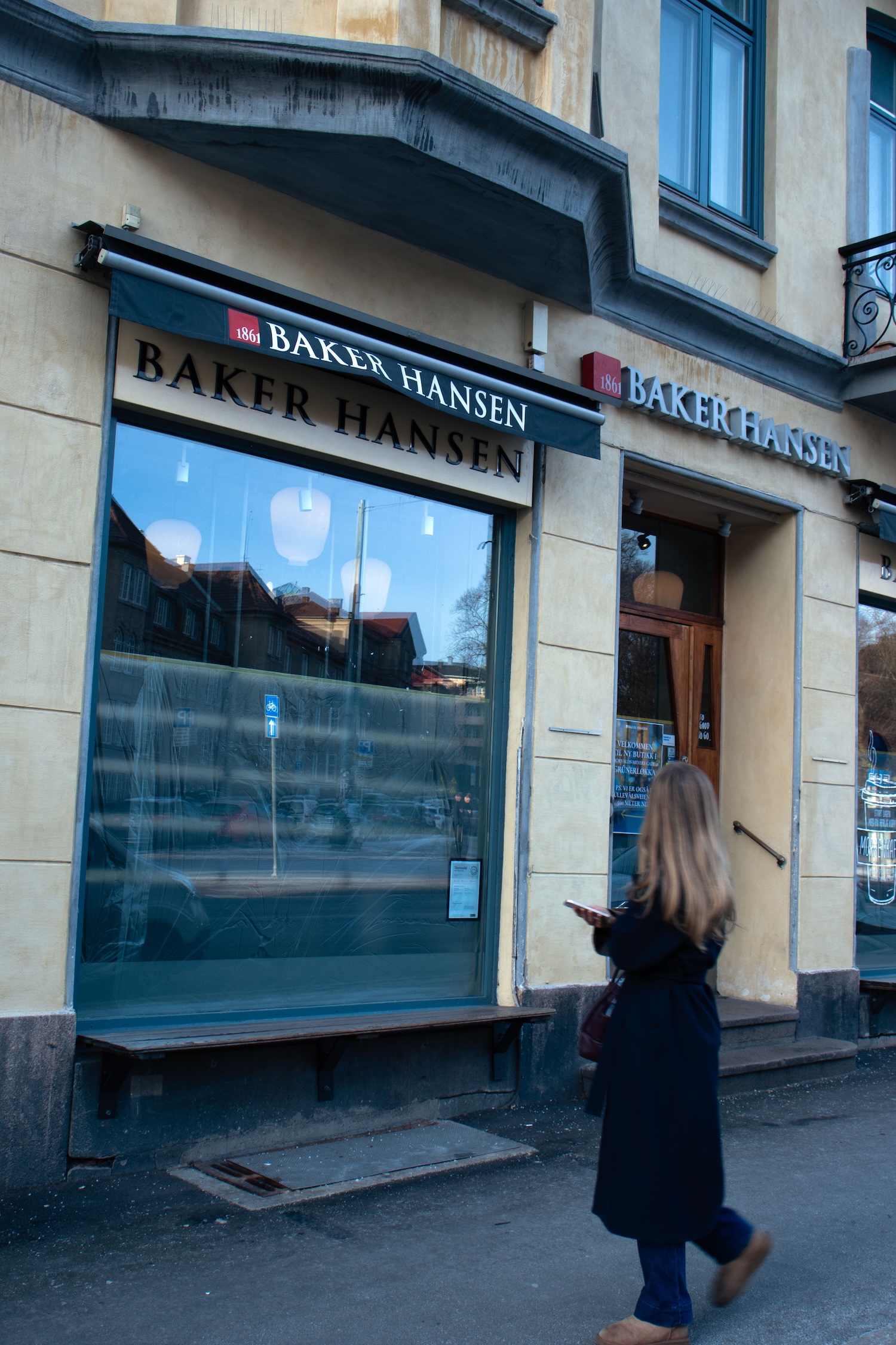 Bildet viser fasaden til Baker Hansen med store blåtonede vinduer og skilt over inngangen, mens en kvinne passerer forbi på fortauet og leser skiltet. 