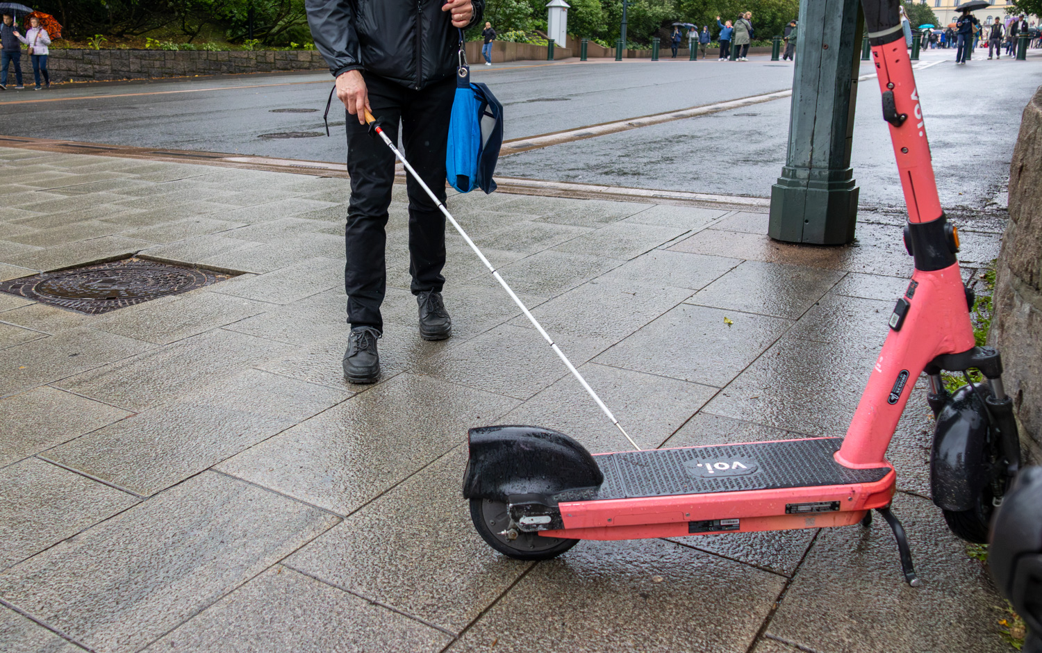 På et fortau, står det en elsparkesykkel. Den står parkert nesten vinkelrett ut fra en vegg. En person, som bare er synlig fra midjen ned, går i retning mot elsparkesykkelen med hvitt stokk i hånd.