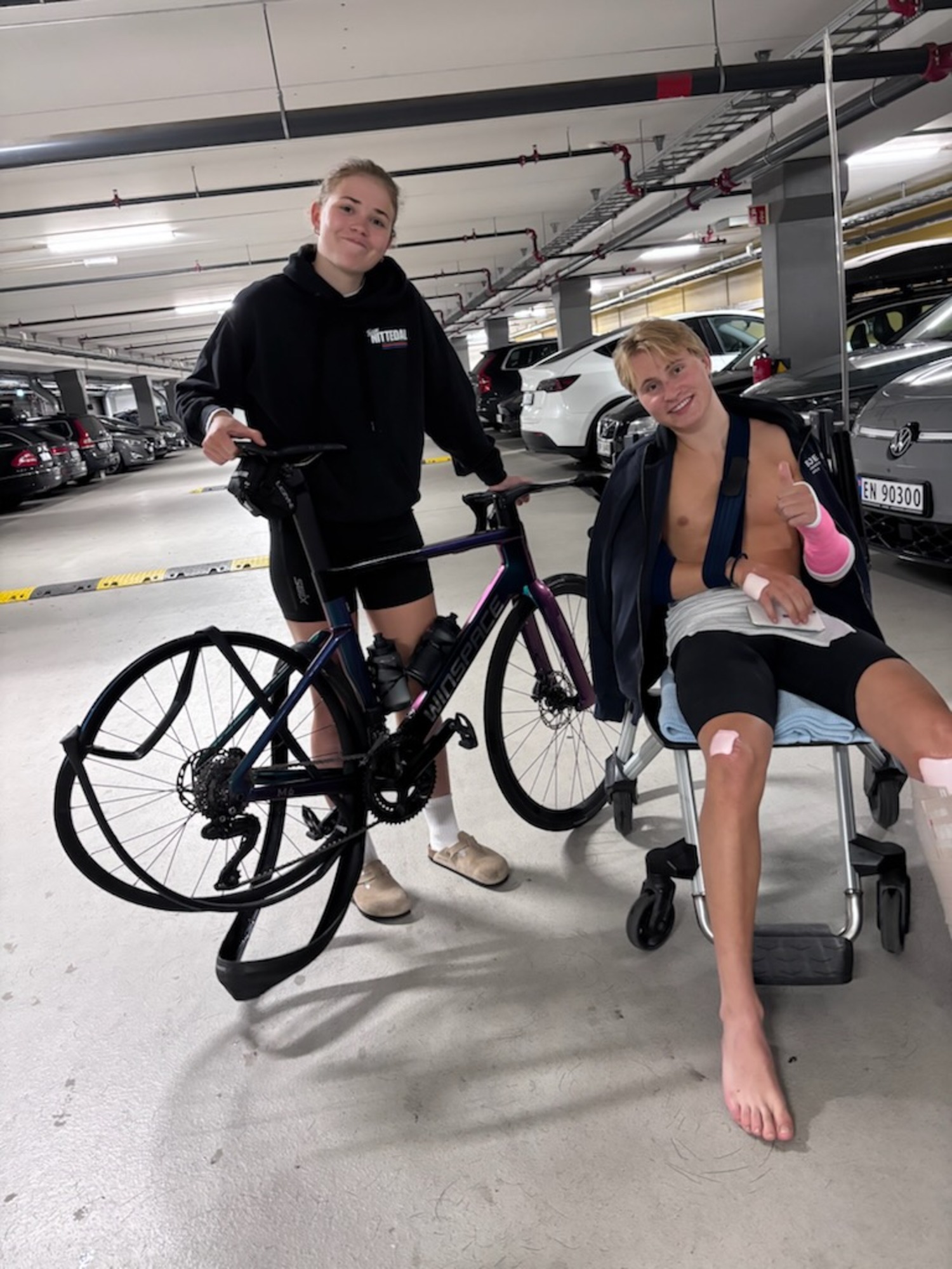 Ei jente holder en ødelagt sykkel opp og en gutt i rullestol sitter ved siden av