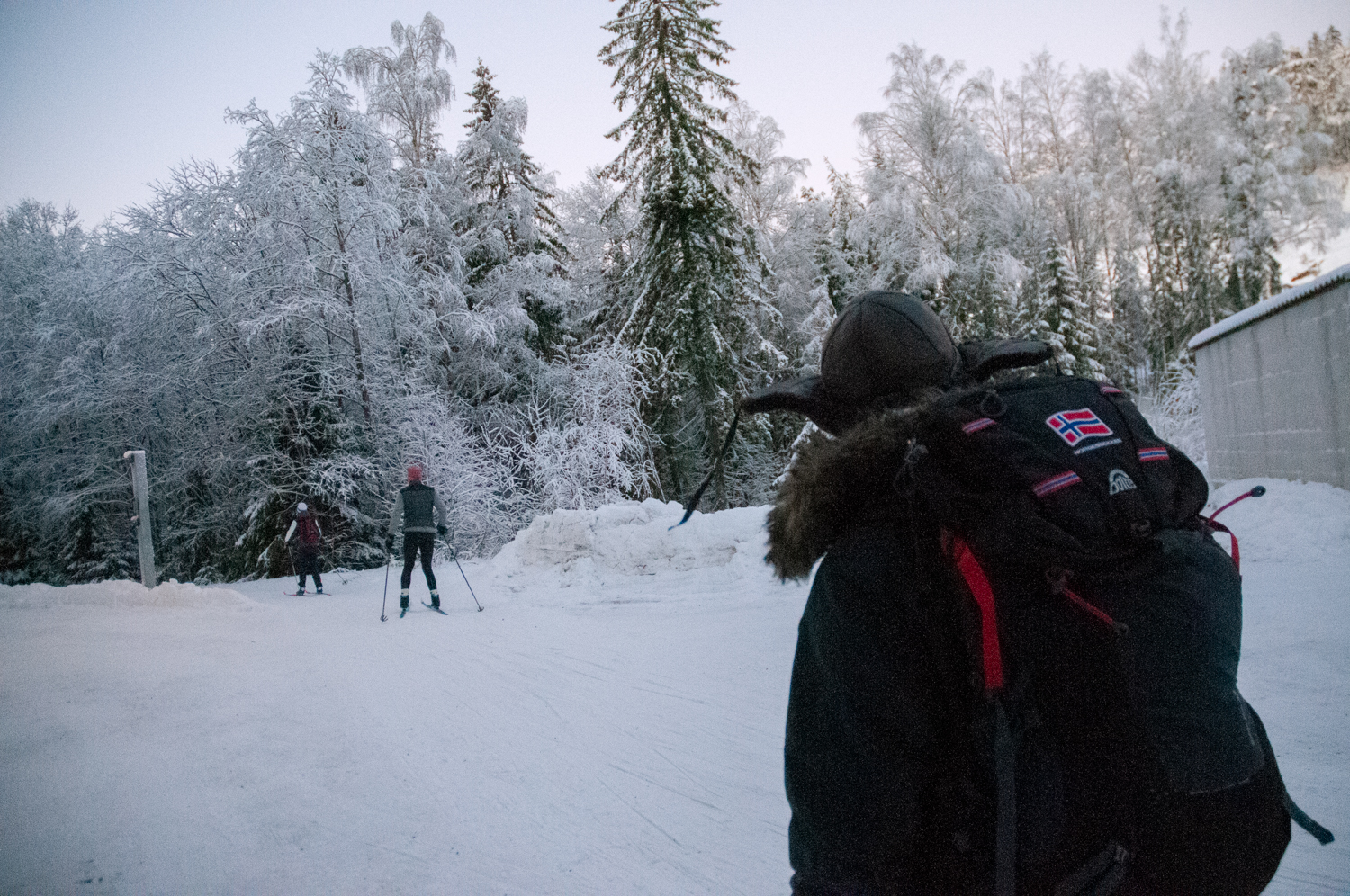 Ryggsekken til Stian imens han går bortover skisporet. Foran han går to skiløpere.
