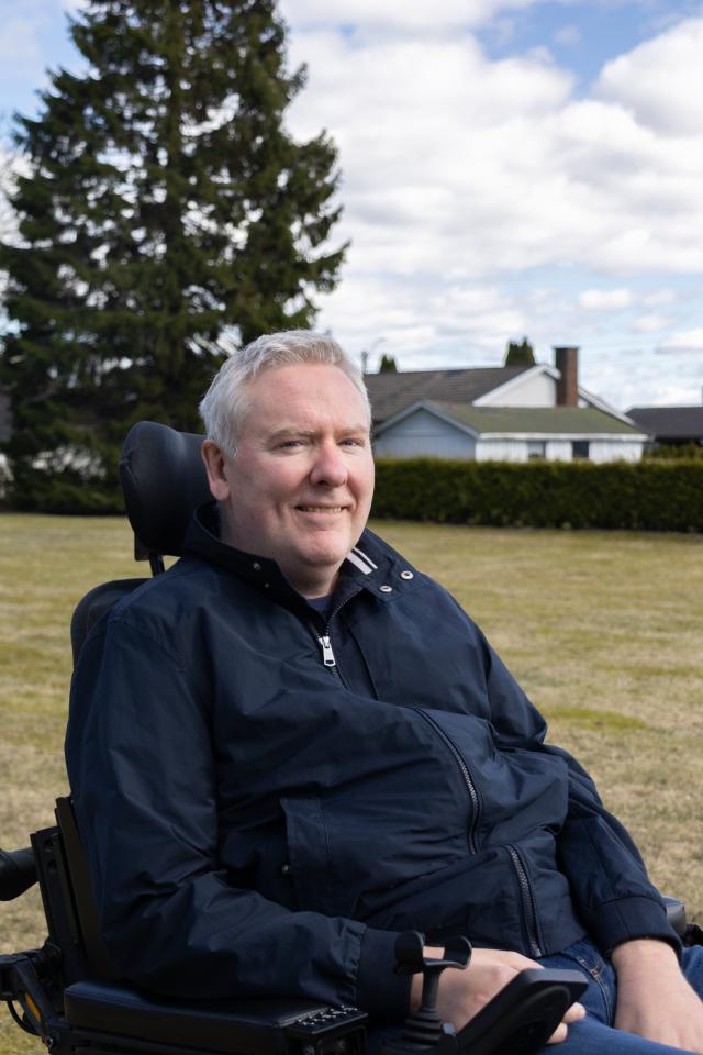 Portrett av Christer. En mann i 50-&aring;ra med gr&aring;tt h&aring;r sitter i rullestol. M&oslash;rkebl&aring; jakke. Gress, tre og hus i bakgrunnen.