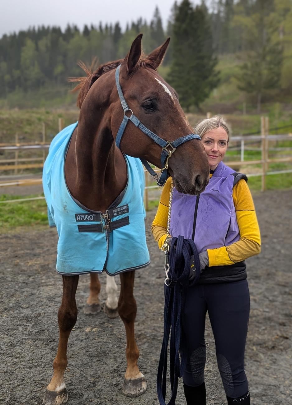 Tina Eriksen og Dayak Z. En blond kvinne med rideklær står ved siden av en brun hest med blått dekken ute på et jorde.