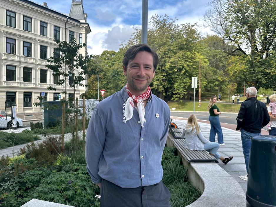Andreas Sjalg Unneland (SV) står utendørs på St. Hanshaugen i Oslo, iført blå skjorte og rødrutete Palestinaskjerf. Bak ham sitter og går folk i parken en solfylt dag.