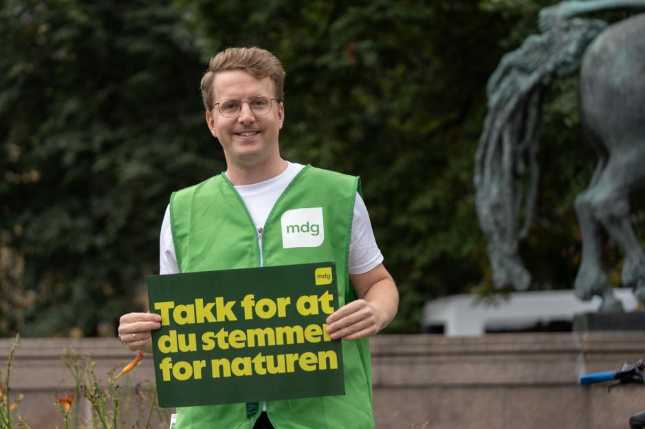 En mann iført grønn vest med MDG logo på, står med ett skilt hvor det står "Takk for at du stemmer for naturen"