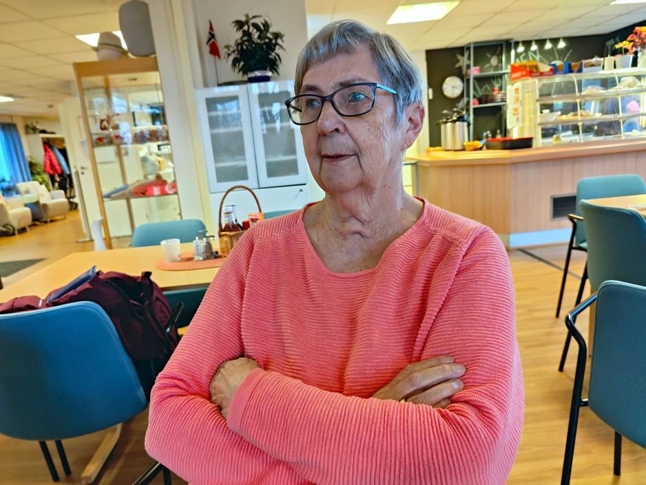 Dame i rosa genser med armene krysset. Briller. Blå stoler bak. 