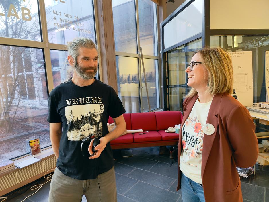 Mann og dame. Mann med sort t-skjorte. Dame med hvit t-skjorte og rød cardigan. Store vinduer bak.