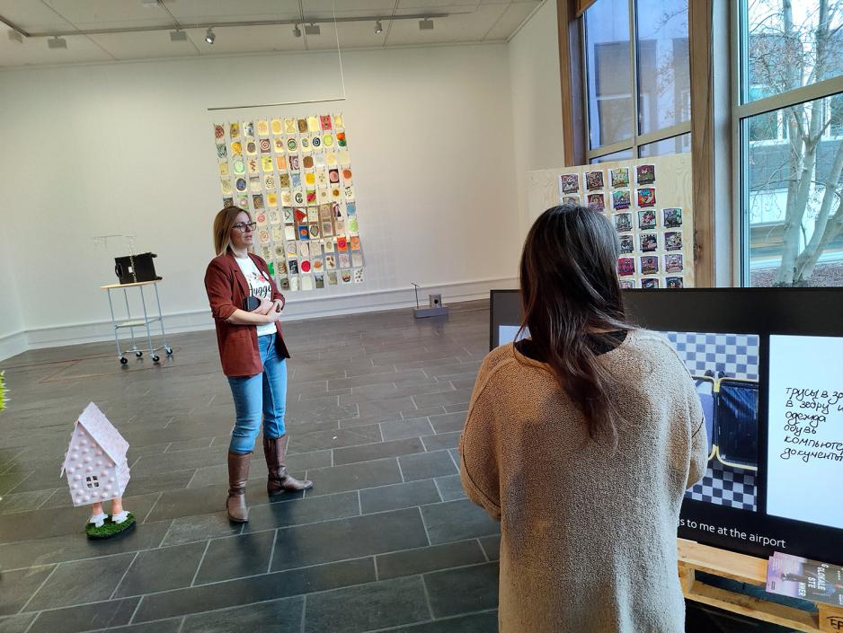 Kunstgalleri. Dame i midten med olabukse og rød blazer. Ser ryggen til dame med langt brunt hår. 