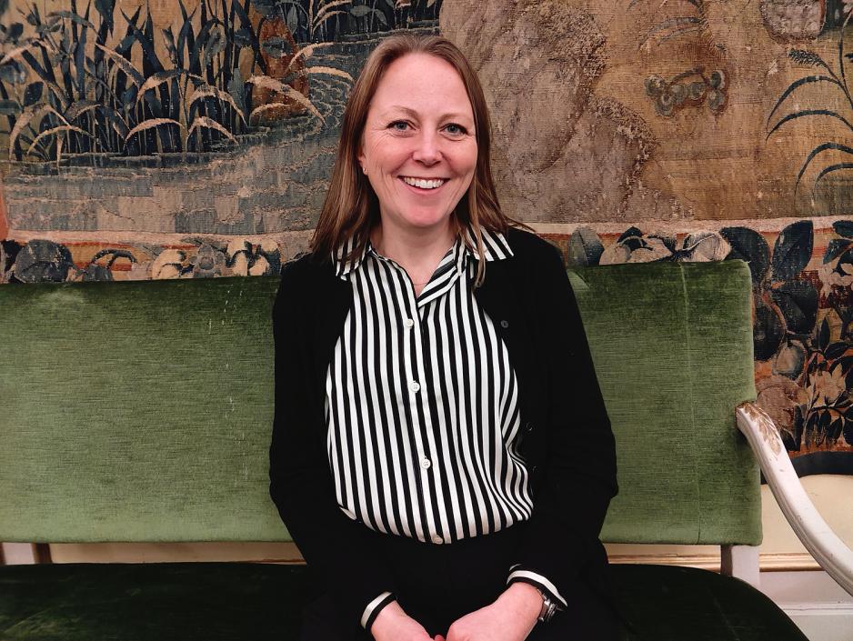 Dame smiler. Stripete skjorte og sort cardigan. Grønn sofa. Maleri i bakgrunn. 