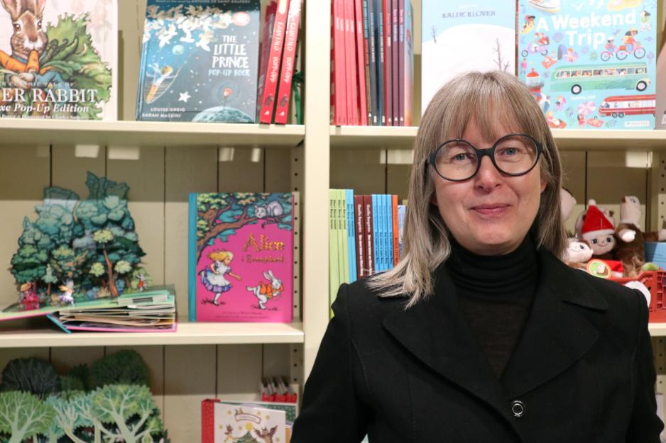Bilde av en dame med briller foran en bokhylle med barnebøker.