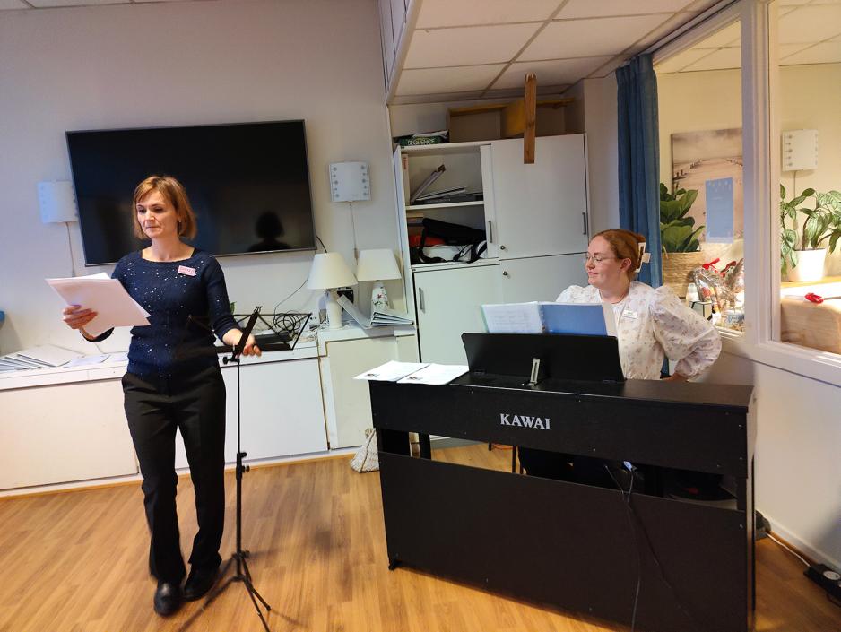 Dame som ser ned på sangtekst. Dame bak sort piano.