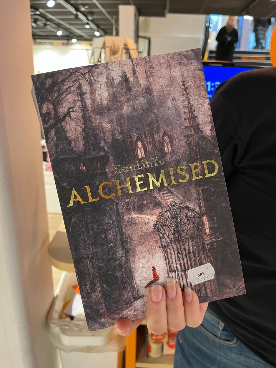 En h&aring;nd holder boka Alchemised av Sen Lin Yu.