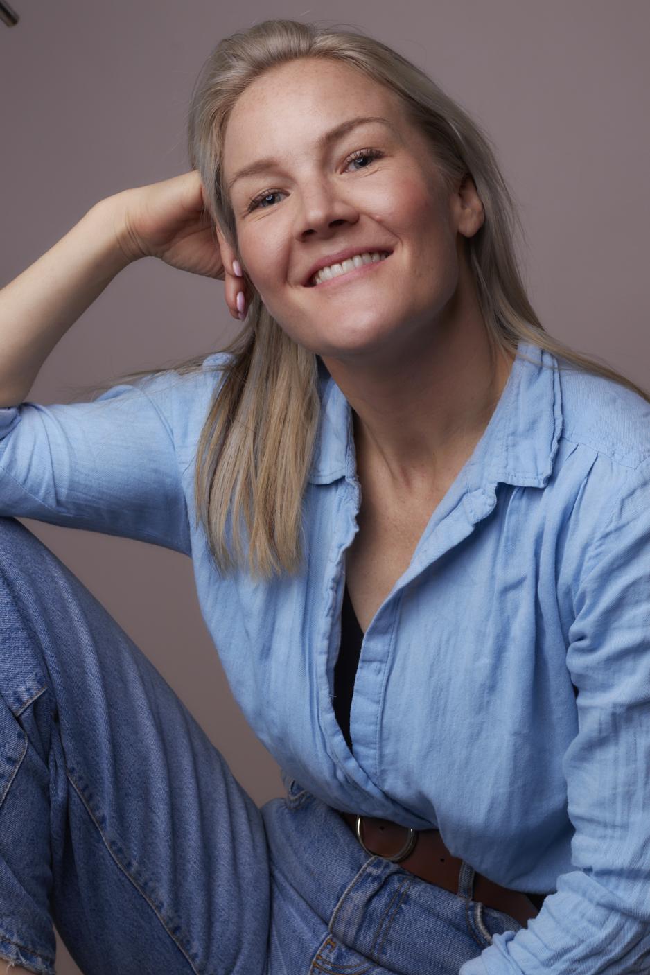 Ida Holten Wors&oslash;e smiler til kamera i bl&aring; skjorte og jeans foran en rosa vegg. hun lener albuen p&aring; kneet og lener hodet mot h&aring;nden