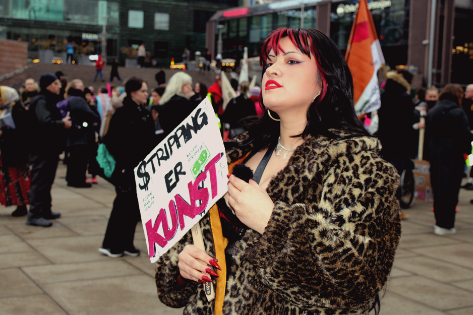 Stripper Alma Adoreholder en plakat der det st&aring;r "Stripping er Kunst". Med haka rettet oppover ser hun bestemt ut