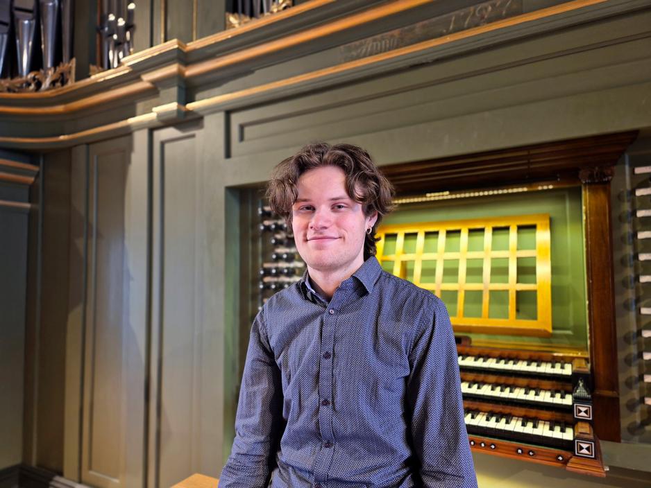 En ung mann i bl&aring; skjorte sitter foran et orgel og smiler mot kamera.