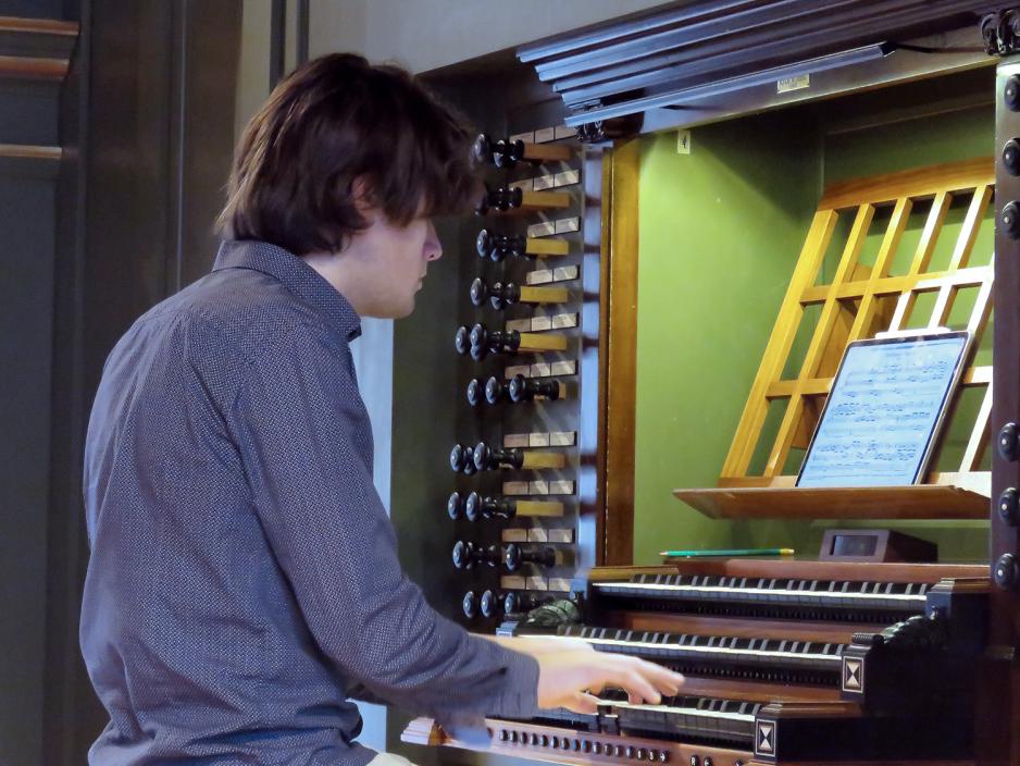 En ung mann i bl&aring; skjorte sitter med ryggen til og spiller orgel. Bildet er tatt fra en sidevinkel.