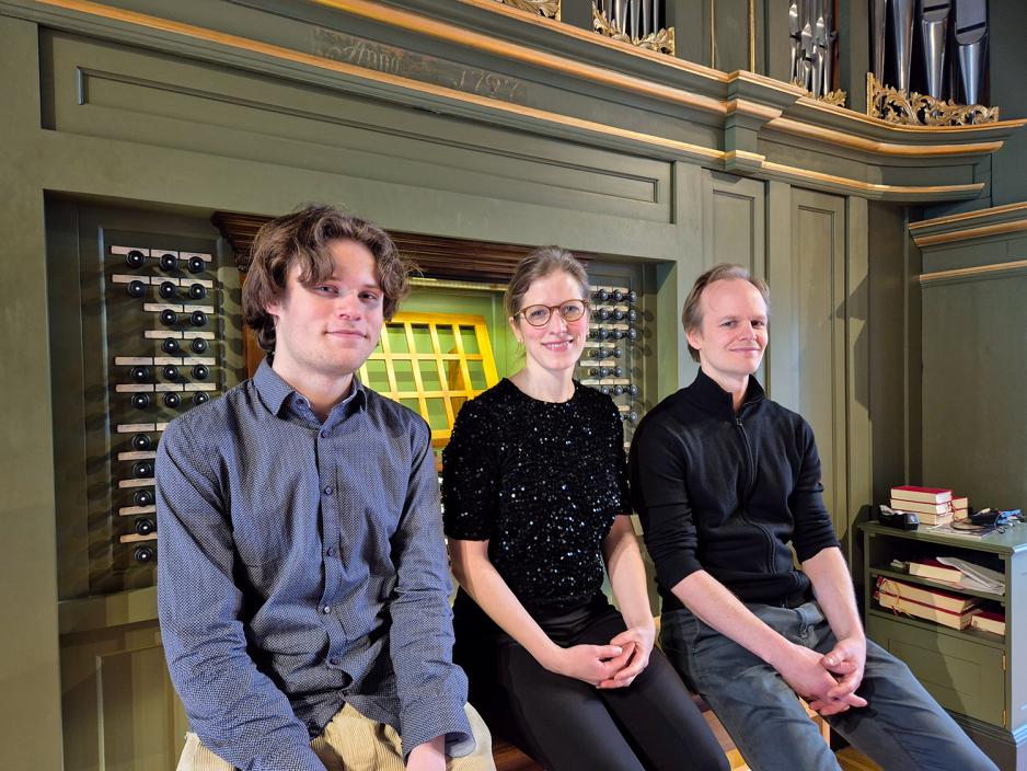 To menn sitter foran et orgel med en kvinne sittende mellom dem. De smiler mot kamera.