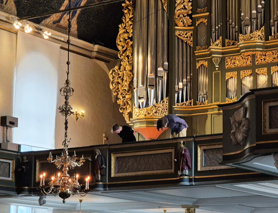 En dame og mann st&aring;r oppe i galleriet i Oslo domkirke, foran hovedorgelet. De bukker ned til et publikum som ikke er med i bildet.