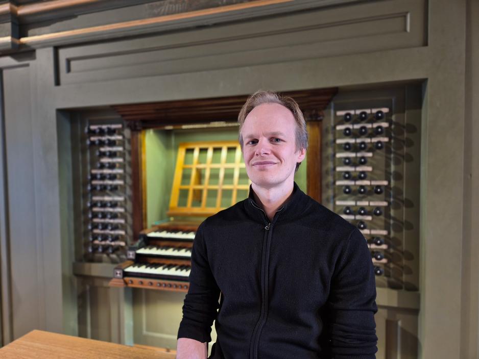 En mann i svart skjorte sitter foran et orgel. Han smiler mot kameraet.