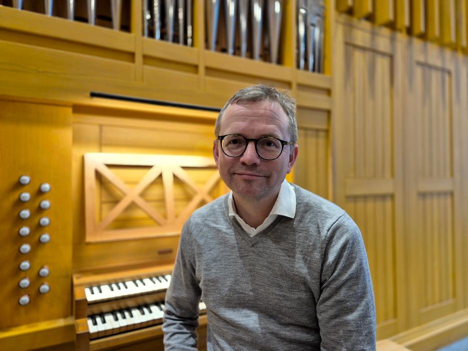 En mann i gr&aring; genser og sorte briller sitter foran et orgel.