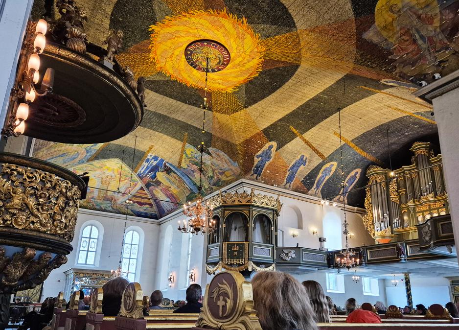 Bilde av taket til Oslo domkirke, med fargerike motiver. Nederst i bildet kan man se hodene til publikum.