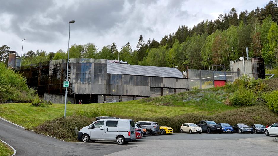 Et moderne bygg ligger p&aring; en liten h&oslash;yde med en parkeringsplass litt i forgrunnen. I bakgrunnen er det gr&oslash;nn skog.