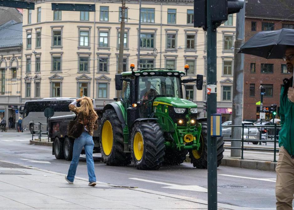 En traktor kjører ned veien på Majorstua. Det er lettregn, og i forgrunnen går en ung jente på gata mens hun prøver å beskytte håret fra regnet.