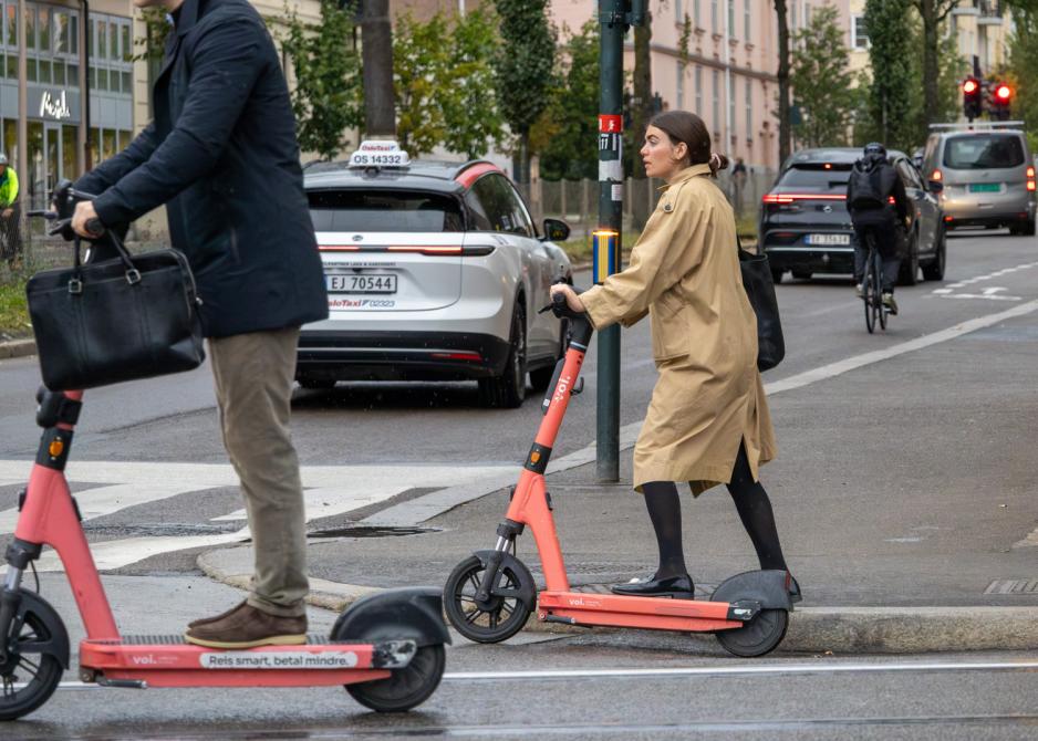 Hovedmotivet er en ung dame på en elsparkesykkel. Hun har en lang kåpe på, og venter på sin tur for å kjøre ut i krysset. I forgrunnen passeres hun av en mann på en elsparkesykkel. Fjeset hans er ikke synlig, men han har på en kort frakk, og holder en PC-bag i venstre hånd. Begge hendene er på rattet.