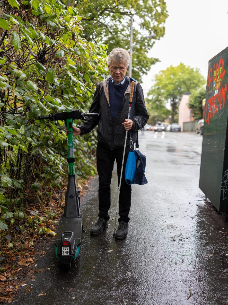 En mann med hvit stokk står på et trangt fortau, ved siden av en elsparkesykkel som er parkert til siden. Til tross for at den er til siden, så er den et hinder. Mannen holder stokken i hans venstre hånd, mens den andre hånden holder elsparkesykkelen under rattet. Han har en PC-bag på hans venstre skulder.