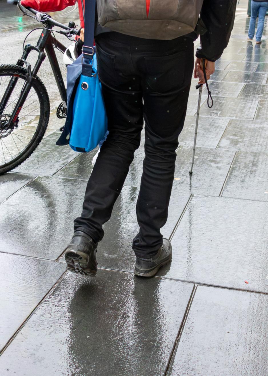 En mann er avbildet bakfra, fra midjen ned. Han har på seg en sekk og en bag på venstre skulder, og holder en stokk med høyre hånd. Han beveger seg bortover på fortauet, og er i ferd med å passere en syklist som går ved siden av sykkelen sin. Fortauet er vått.