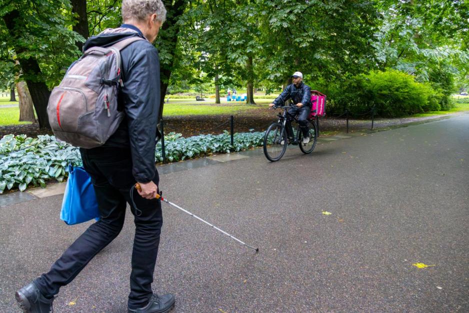 En mann med hvit stokk i høyre hånd går gjennom en park. Bildet er tatt fra en sidevinkel, litt bak han på hans høreside. Han har på en stor sekk. En syklist kommer mot han på andre halvdel av veien.