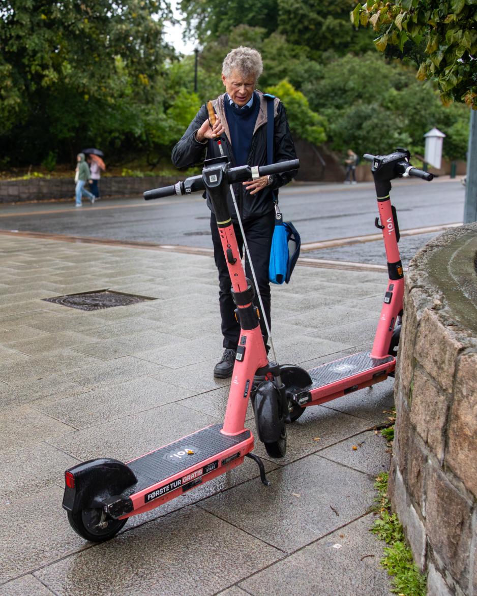 En svaksynt mann med stokk står bak to elsparkesykler på et bredt fortau. Elsparkesyklene er parkert slik at de fortsatt stikker ut i gåarealet. 