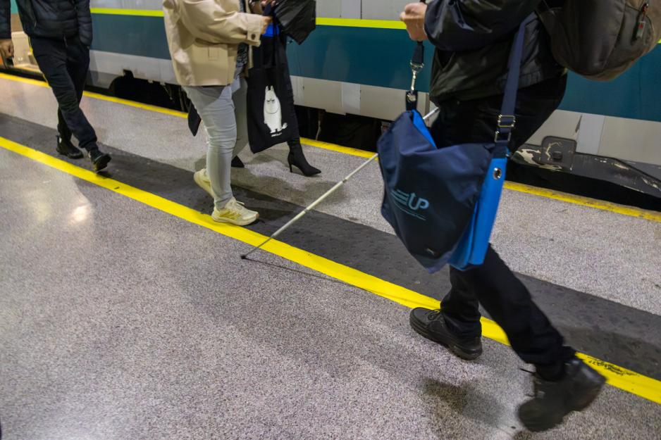 Bildet viser noen med stokk som går på en tog plattform. Vedkommende har på seg en sekk og en bag, og følger ledelinjene, hvor flere folk står i veien. Vedkommende er kun avbildet fra midjen ned.