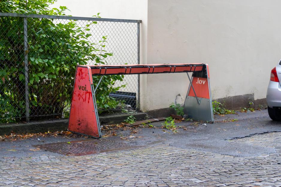 Bildet viser et stativ hvor man kan parkere voi-elsparkesykler. Den står på gata bak en bil, er tom og full av grafitti. 