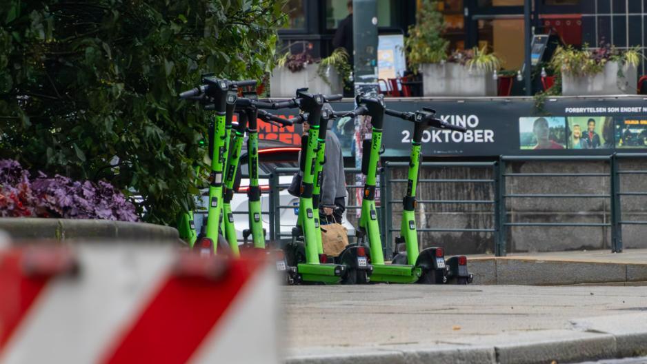 Bildet er tatt fra en avstand. Hovedmotivet i midten av bildet er en haug av elsparkesykler som er parkert sammen på et svinget fortau.