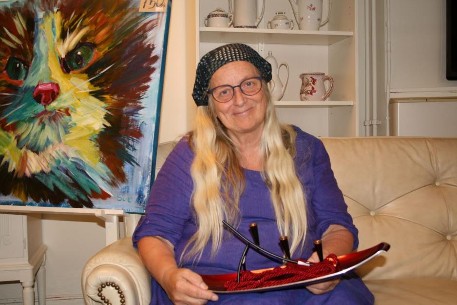 Lisbeth Wood sitter i en sofa med lilla kjole og skaut. Hun holder noen varer i hendene og smiler.